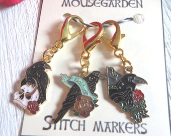 May include: Three gold stitch markers with black raven charms. The charms feature different designs, including a skull, a rose, and a banner with the word "Solitude".