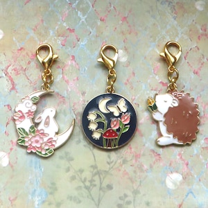 以下が含まれることがあります： エナメルデザインの金色のチャームクリップ3つ。最初のチャームは、ピンクの花が咲いた三日月に座っている白いウサギ。2つ目のチャームは、三日月、白い花、赤いキノコ、蝶が描かれた青い円形。3つ目のチャームは、黄色い花を持った茶色のハリネズミです。
