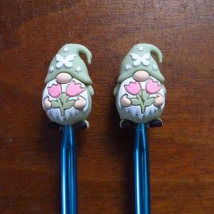May include: Two pens with gnome toppers. The gnomes have green hats with white butterfly and star accents, beige noses, and white beards. They hold pink tulips. The pens have blue metallic barrels and black feet.
