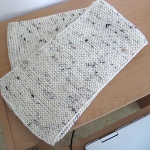May include: A cream-colored, hand-knitted scarf with a speckled pattern of black and brown flecks. The scarf is folded over itself, displaying the textured knit. The scarf is made of a soft yarn and is suitable for cold weather.