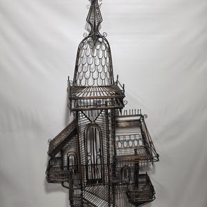 May include: A large, dark brown metal birdcage featuring an intricate, multi-level architectural design with a tall, pointed spire at the top, arched doorways, tiered balconies, and a complex wire framework that resembles a miniature castle structure.
