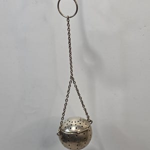 May include: A spherical metal tea infuser with a perforated surface, featuring a hinged opening mechanism. The infuser is suspended by two thin metal chains that connect to a single circular metal ring at the top for hanging or holding.