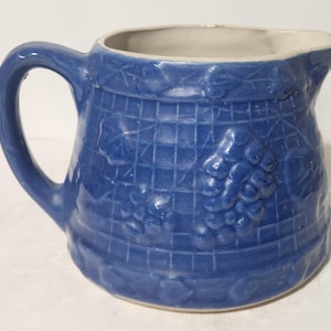 May include: A blue ceramic pitcher with a handle and spout. The pitcher has a textured surface with a grid pattern and grape cluster design. The interior is white.