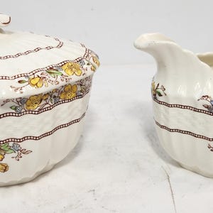 May include: Cream-colored ceramic sugar bowl with lid and creamer. Both pieces feature a floral design with yellow and blue flowers, brown trim, and a ribbed texture. The sugar bowl has a decorative handle on the lid.