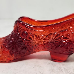 May include: A decorative, ruby-red glass shoe with a textured, starburst pattern. The vintage-style shoe features a small heel and a detailed design, perfect for display or as a unique accent piece.
