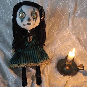 May include: A handcrafted fabric doll with long black yarn hair, button eyes, and a pale face featuring teal and black painted details. The doll wears a green and black striped dress with gold lace trim and a small cameo pendant at the neck.