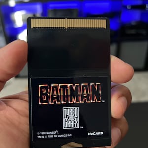 May include: Black HuCARD video game cartridge for the TurboGrafx-16. The cartridge features the Batman logo in orange and yellow, with the TurboGrafx-16 logo below. The text "&copy; 1990 SUNSOFT. TM & &copy; 1989 DC COMICS INC." is also visible.