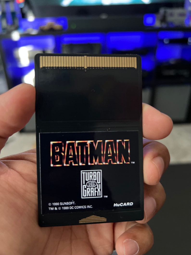 May include: Black HuCARD video game cartridge for the TurboGrafx-16. The cartridge features the Batman logo in orange and yellow, with the TurboGrafx-16 logo below. The text "&copy; 1990 SUNSOFT. TM & &copy; 1989 DC COMICS INC." is also visible.