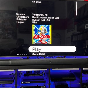 May include: A television screen displays the game "Air Zonk" for the TurboGrafx-16 system. The game's title is in red and yellow, with a cartoon character. The screen shows "Play" and "Game Detail" options. A black soundbar sits below the screen.
