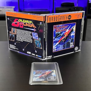 May include: A TurboGrafx-16 video game case for Soldier Blade, with the game cartridge in front. The case is open, displaying the game's cover art with a red and white spaceship design. The text "SOLDIER BLADE" is visible.