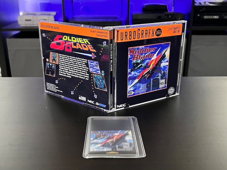 May include: A TurboGrafx-16 video game case for Soldier Blade, with the game cartridge in front. The case is open, displaying the game's cover art with a red and white spaceship design. The text "SOLDIER BLADE" is visible.