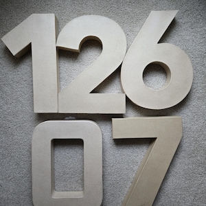 May include: A collection of large, beige cardboard numbers: 1, 2, 6, 0, and 7. The numbers are arranged on a gray surface. The numbers are thick and three-dimensional, suitable for crafting or decorative purposes.