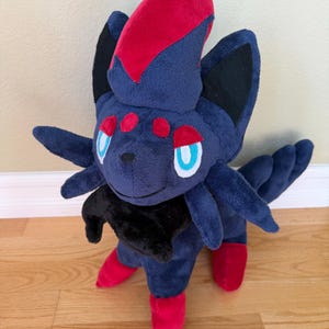 May include: A plush toy of a dark blue creature with red accents. The toy has a red crest, red feet, and red spots. It has black ears and a black collar. The eyes are blue with white outlines. The plush is standing on a wooden floor.
