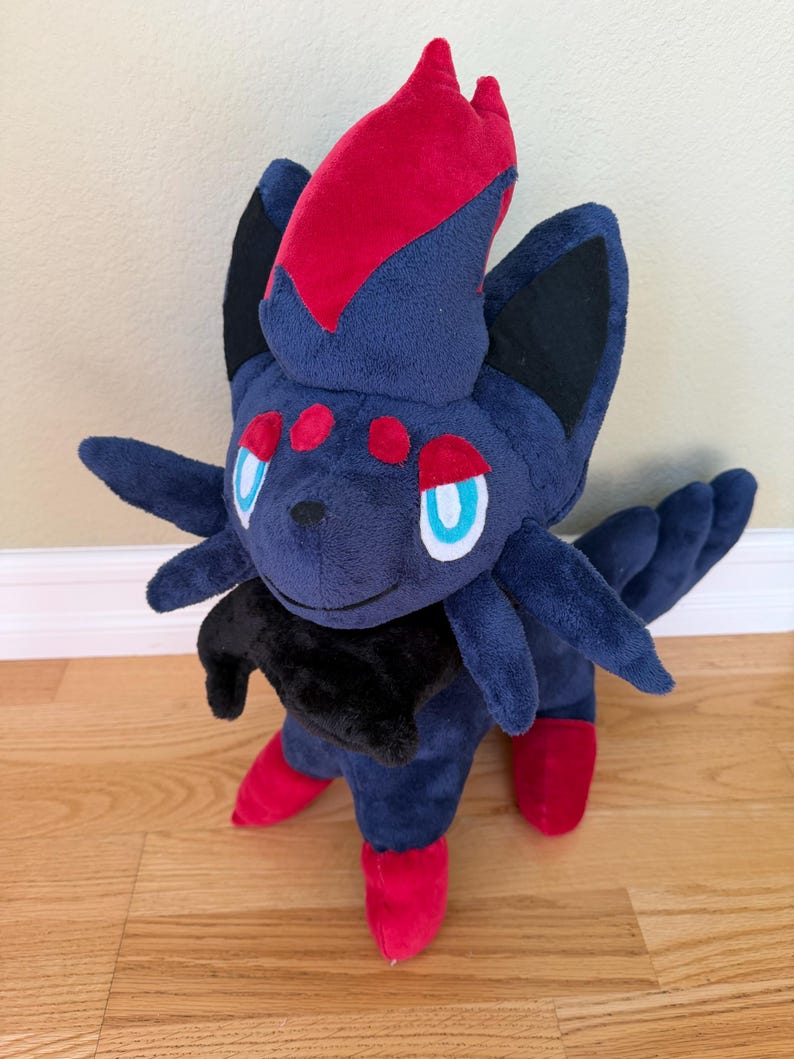 May include: A plush toy of a dark blue creature with red accents. The toy has a red crest, red feet, and red spots. It has black ears and a black collar. The eyes are blue with white outlines. The plush is standing on a wooden floor.