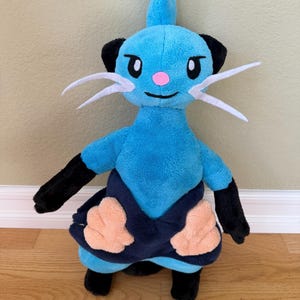 May include: A plush toy of a blue creature with black accents, white whiskers, and a pink nose. The toy has black arms and legs, and a dark blue skirt with peach-colored details. The toy is standing on a wooden floor.