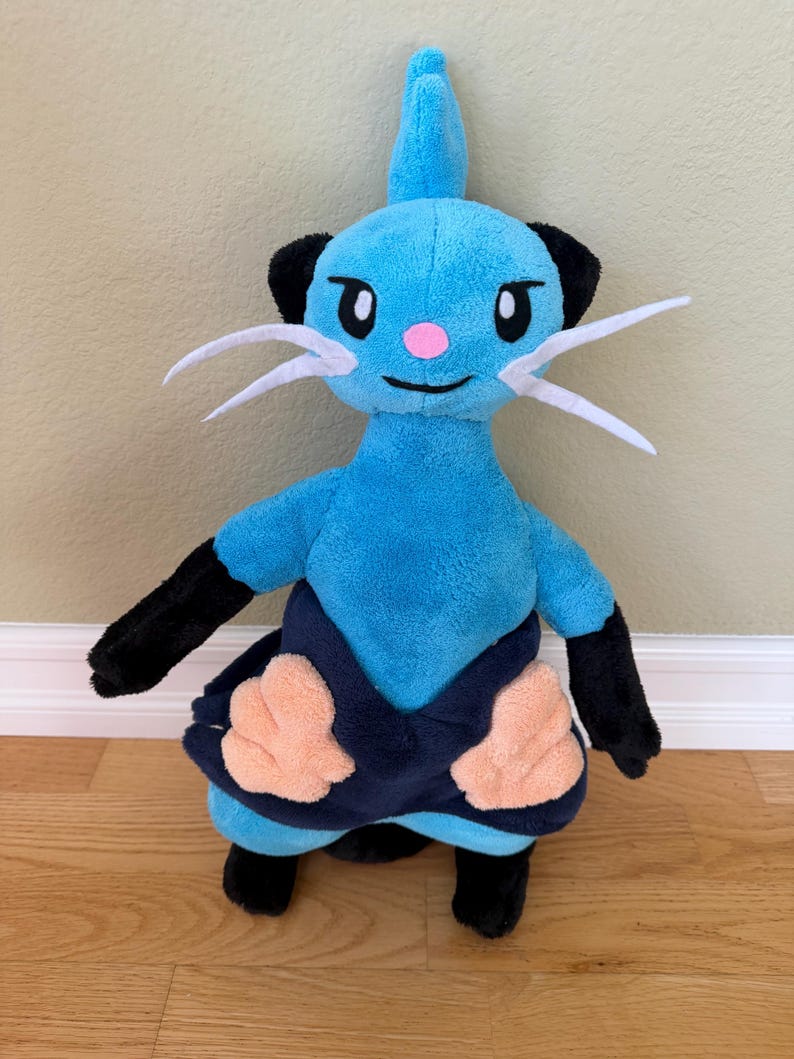 May include: A plush toy of a blue creature with black accents, white whiskers, and a pink nose. The toy has black arms and legs, and a dark blue skirt with peach-colored details. The toy is standing on a wooden floor.
