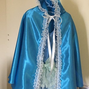 May include: A bright blue satin cape with a hood, trimmed with white lace. A white ribbon ties the cape at the neck. The cape is part of a costume, likely for a child.