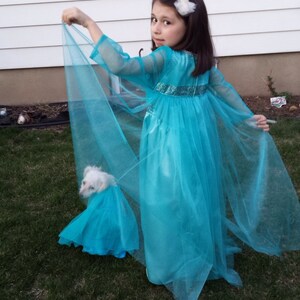 Puede incluir: Una ni&ntilde;a peque&ntilde;a con un vestido turquesa con mangas transparentes y una capa a juego. Un perro blanco peque&ntilde;o tambi&eacute;n lleva un vestido turquesa a juego. La ni&ntilde;a est&aacute; de pie sobre hierba verde.