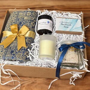 May include: A gift box filled with a blue and gold patterned scarf, a jar of bath salts with a blue ribbon, a candle in a frosted glass jar, a bar of soap with the text "All Natural Handmade Soap", and a small jar of moisturizer.