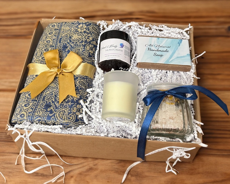 May include: A gift box filled with a blue and gold patterned scarf, a jar of bath salts with a blue ribbon, a candle in a frosted glass jar, a bar of soap with the text "All Natural Handmade Soap", and a small jar of moisturizer.