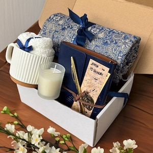 May include: A white gift box filled with a blue and white patterned throw blanket, a blue journal with a gold pen, a white candle, and a white mug with a blue ribbon.