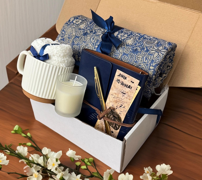 May include: A white gift box filled with a blue and white patterned throw blanket, a blue journal with a gold pen, a white candle, and a white mug with a blue ribbon.
