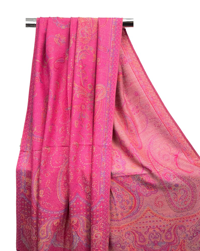 May include: A vibrant fuchsia pink shawl featuring an intricate woven paisley pattern in shades of orange, purple, and light pink. The fabric displays a dense, textured design with a wide decorative border along the edge, draped over a metal rod.
