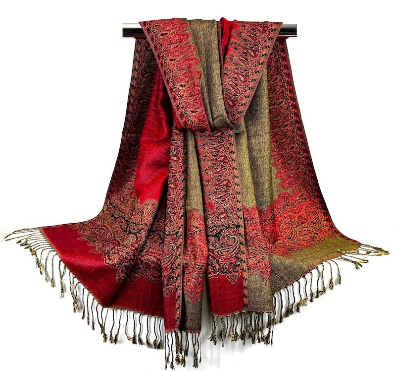 Pode incluir: Um xale vermelho e verde com padr&atilde;o paisley e franjas douradas. O xale est&aacute; dobrado ao meio e pendurado em um cabide.
