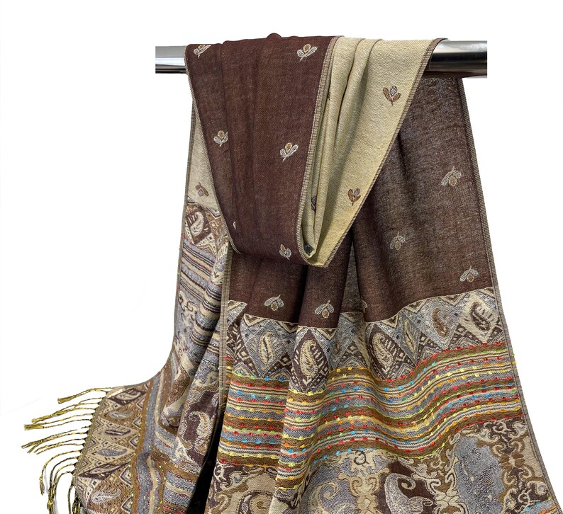 K&ouml;nnte beinhalten: Ein brauner und beiger Schal mit einem detaillierten Paisley- und Streifenmuster. Der Schal hat am Ende Fransen und ist &uuml;ber einer Metallstange drapiert. Der Schal hat ein reversibles Design mit einer einfarbigen braunen und einer beigen Seite.