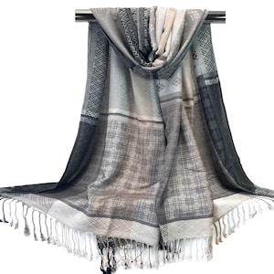 May include: A gray and white scarf with a geometric pattern and fringe. The scarf is draped over a metal bar, showcasing its design. The color palette includes shades of gray, white, and black, creating a neutral and versatile accessory.