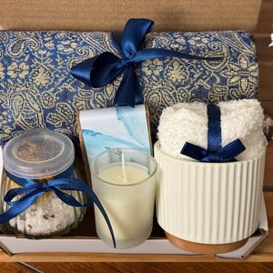 May include: A gift basket with a blue and gold patterned throw blanket, a white ceramic candle holder with a blue ribbon, a glass jar with bath salts, a white candle, and a bar of soap.