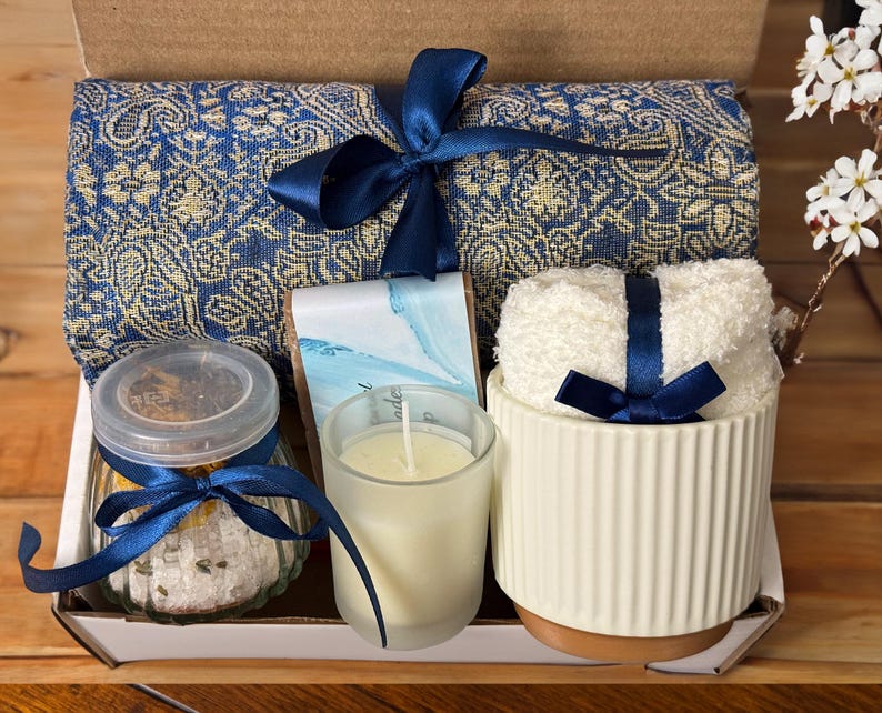 May include: A gift basket with a blue and gold patterned throw blanket, a white ceramic candle holder with a blue ribbon, a glass jar with bath salts, a white candle, and a bar of soap.