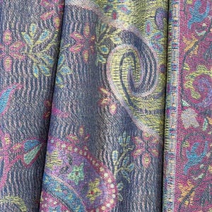 May include: A close-up of a fabric with a paisley pattern in shades of purple, blue, yellow, and pink. The fabric has a textured surface and a woven border.
