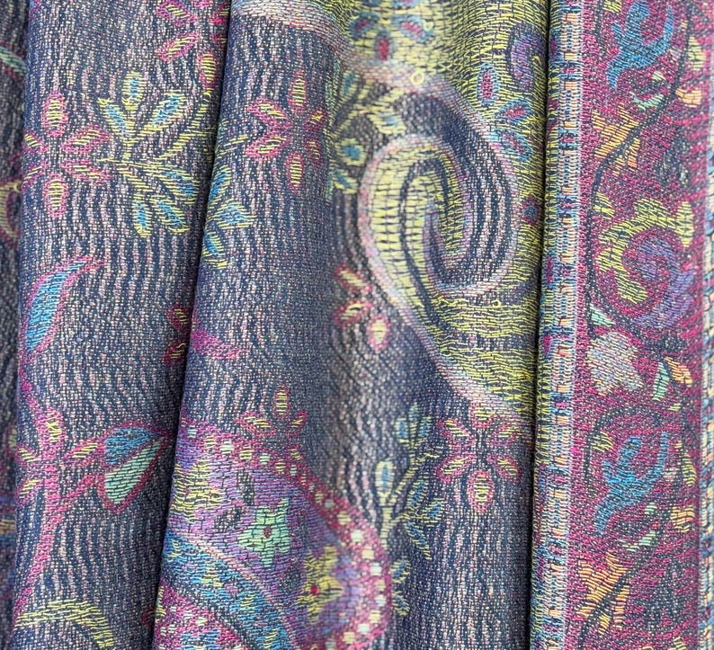 May include: A close-up of a fabric with a paisley pattern in shades of purple, blue, yellow, and pink. The fabric has a textured surface and a woven border.