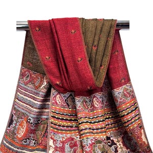 K&ouml;nnte beinhalten: Ein roter und brauner Schal mit detailliertem Paisley-Muster und floralen Akzenten. Der Schal ist &uuml;ber einer Metallstange drapiert und zeigt sein aufwendiges Design und seine Farbpalette. Die Kanten des Schals weisen einen detaillierten Rand auf.