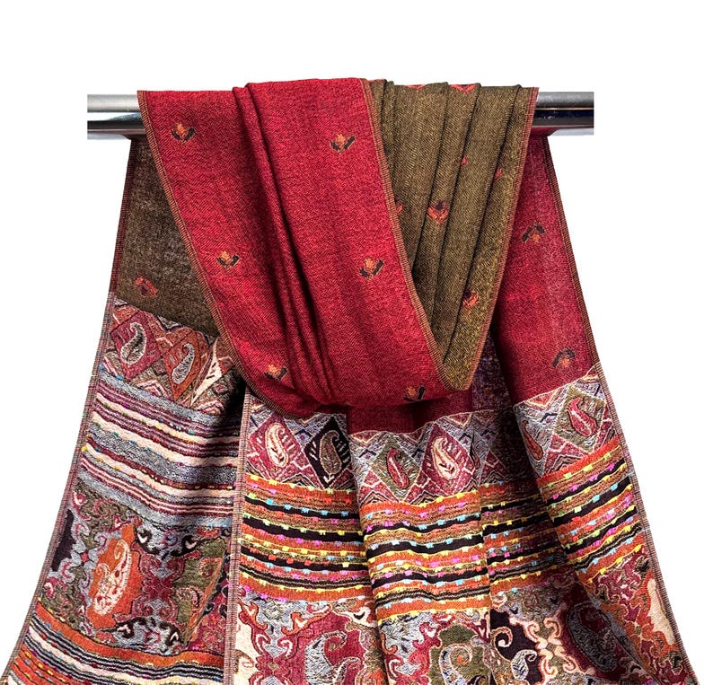K&ouml;nnte beinhalten: Ein roter und brauner Schal mit detailliertem Paisley-Muster und floralen Akzenten. Der Schal ist &uuml;ber einer Metallstange drapiert und zeigt sein aufwendiges Design und seine Farbpalette. Die Kanten des Schals weisen einen detaillierten Rand auf.