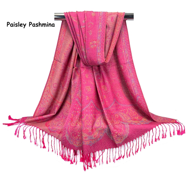 May include: A bright pink pashmina shawl featuring a woven paisley pattern in gold and light blue tones. The fabric has a textured appearance with a decorative border and long, thin fringe tassels hanging from the bottom edge. Text reads Paisley Pashmina.