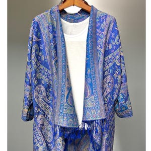 May include: A blue and purple paisley patterned kimono with a fringe trim. The kimono is open in the front and has a white undershirt.