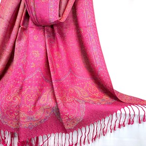May include: A vibrant fuchsia pink scarf featuring an intricate woven paisley pattern in shades of gold and light blue. The rectangular fabric has a textured finish and is finished with long, thin fringe tassels along the bottom edge.