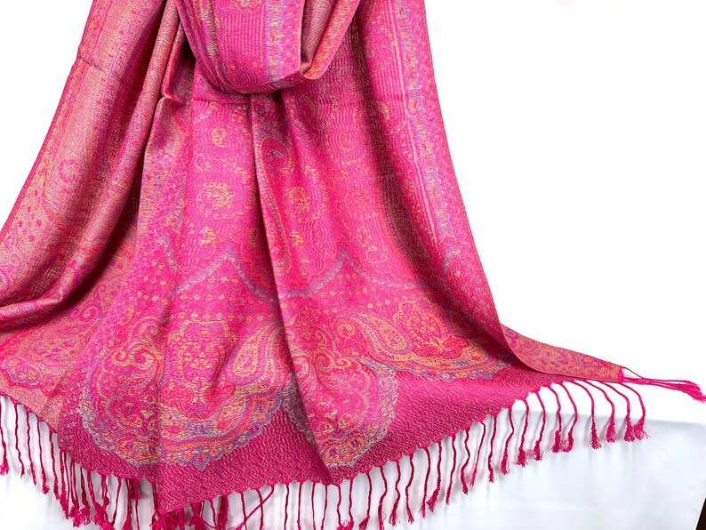 May include: A vibrant fuchsia pink scarf featuring an intricate woven paisley pattern in shades of gold and light blue. The rectangular fabric has a textured finish and is finished with long, thin fringe tassels along the bottom edge.