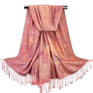 May include: A pink and gold paisley patterned scarf with a fringed edge. The scarf is draped over a metal bar, showcasing its intricate design and color variations. The scarf's design includes floral and paisley motifs.