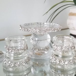 May include: Clear glass candle holders with sparkling rhinestone accents. The set includes a small pedestal cake stand and two smaller candle holders. The candle holders are arranged on a mirrored surface, reflecting the light and adding to the elegant aesthetic.