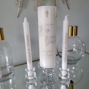 May include: Two white candles with gold lettering on clear glass candle holders. The larger candle has the text "and the two shall become one" and "Michael & Melanie June 17, 2025". The smaller candles have the text "Michael & Melanie".