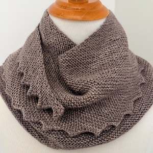May include: A brown knitted cowl scarf featuring a garter stitch texture and a decorative scalloped edge. The scarf is wrapped around a mannequin neck, displaying a soft, uniform yarn construction with a consistent earthy brown color throughout.