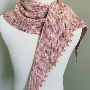 May include: A hand-knitted scarf in a variegated yarn featuring soft shades of pink, lavender, and cream. The scarf is worked in a garter stitch pattern with a distinctively pointed, sawtooth-style edge along one side, creating a textured finish.