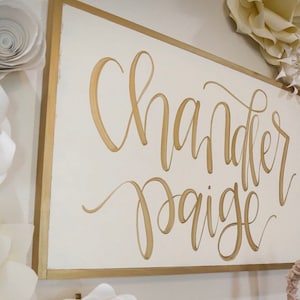 May include: A white sign with a gold frame and gold lettering that reads "Chandler Paige". The sign is surrounded by white paper flowers.