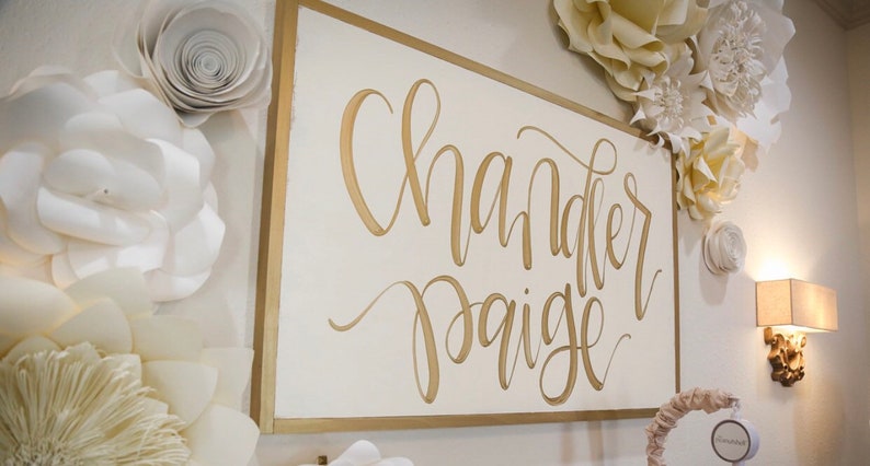 May include: A white sign with a gold frame and gold lettering that reads "Chandler Paige". The sign is surrounded by white paper flowers.