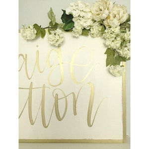 May include: White wooden sign with gold calligraphy that reads "Engagement Station" decorated with white flowers.