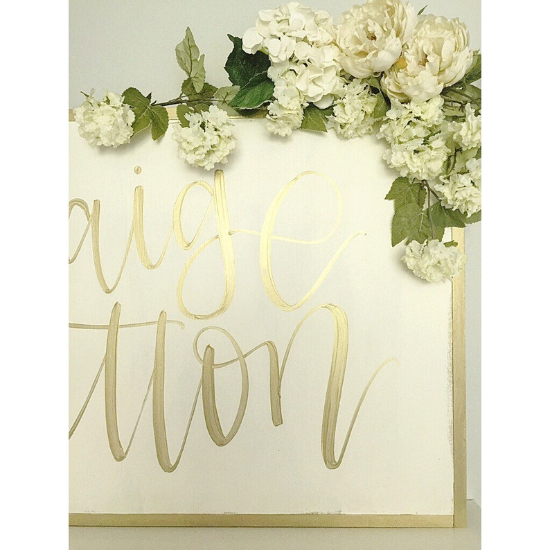 May include: White wooden sign with gold calligraphy that reads "Engagement Station" decorated with white flowers.