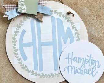 Puede incluir: Un monograma azul claro con las letras "HHM" en una corona de hojas verdes sobre un fondo de cuadros blancos y beige. Un círculo blanco separado con los nombres "Hampton Michael" escritos en azul claro.